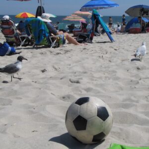 A summer day at the beach with people in chairs under umbrellas. Seagulls are walking along and a soccer ball sits in the foreground