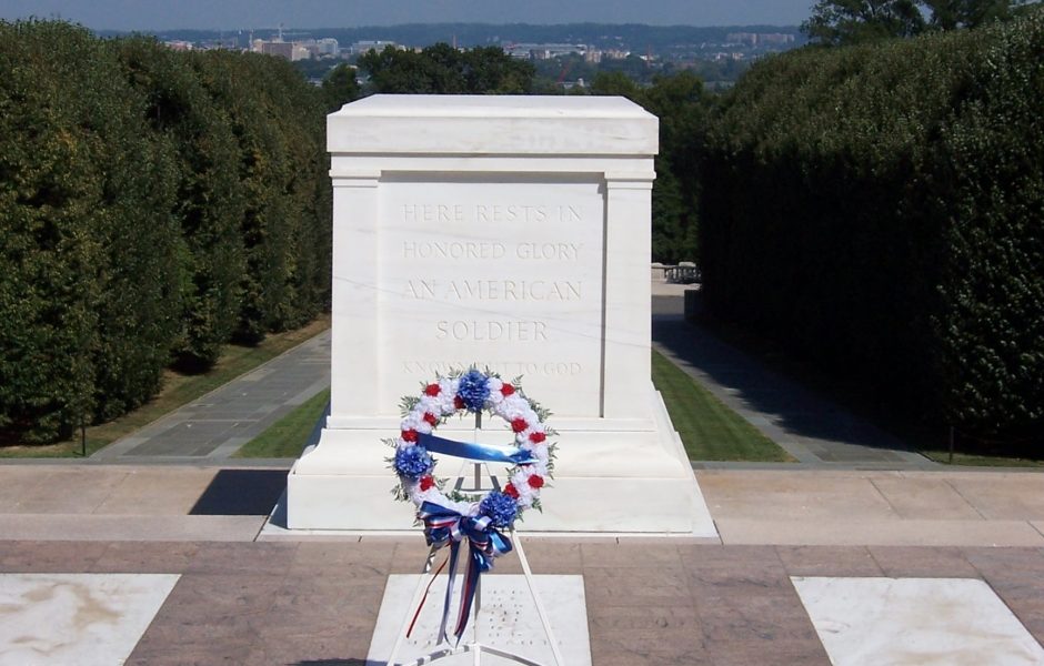 a red, white and blue wreath set on a stnd in front of a memorial to "An American Soldier" with Washington DC in the background