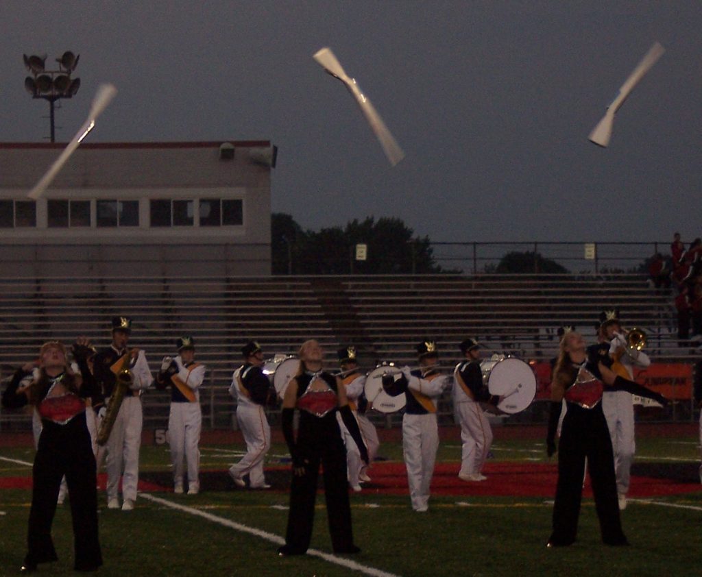 Three girls toss rifles while a marching band performs behind them